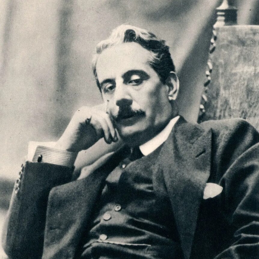 A black and white portrait of Giacomo Puccini sitting in a chair and holding a cigarette.