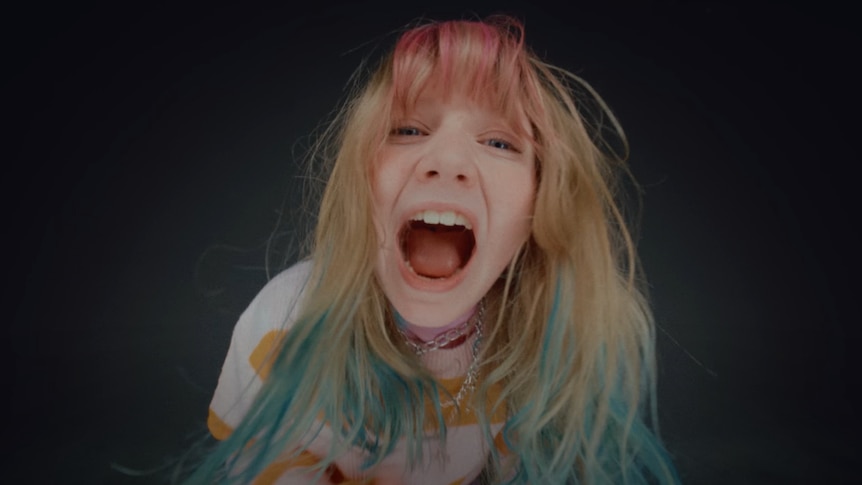 Young vocalist Harper yells at the camera in a white and yellow striped top and pink, blonde and blue hair.