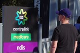 People line up outdoors beside a Centrelink sign.