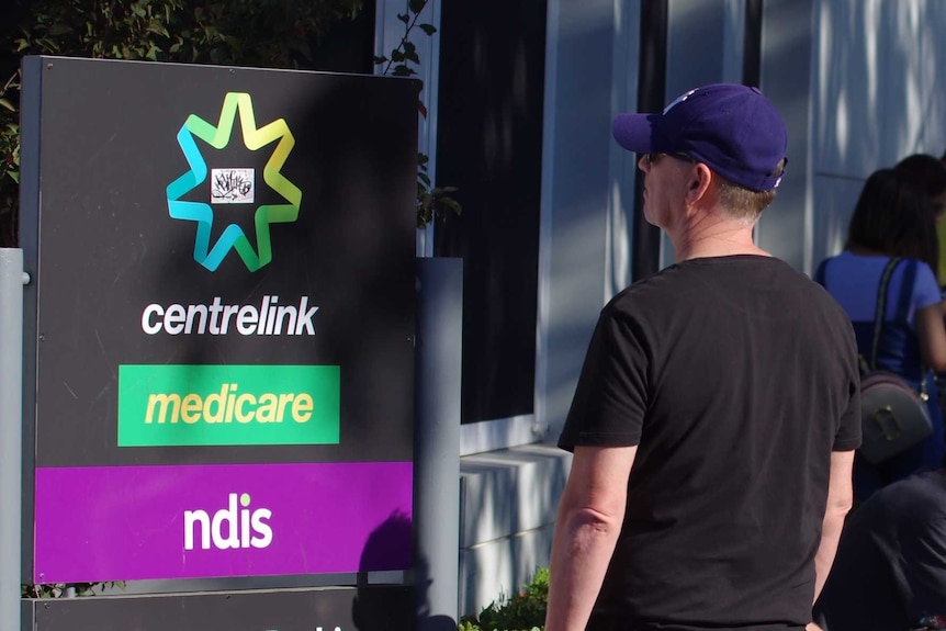 People line up outdoors beside a Centrelink sign.