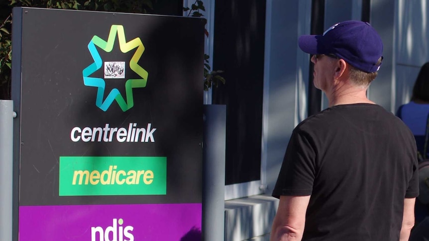 People line up outdoors beside a Centrelink sign.