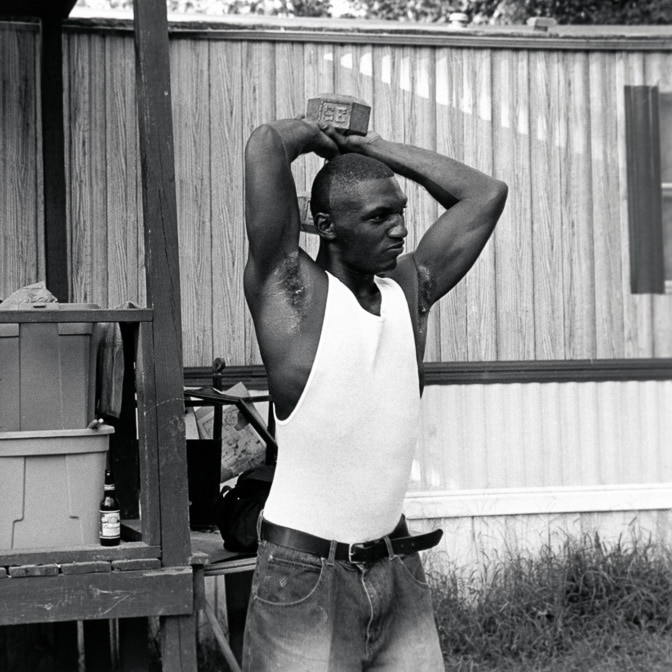 black and white, Cedric is tall and dark skinned, jeans and white singlet, holding a dumbbell, older man on porch behind him