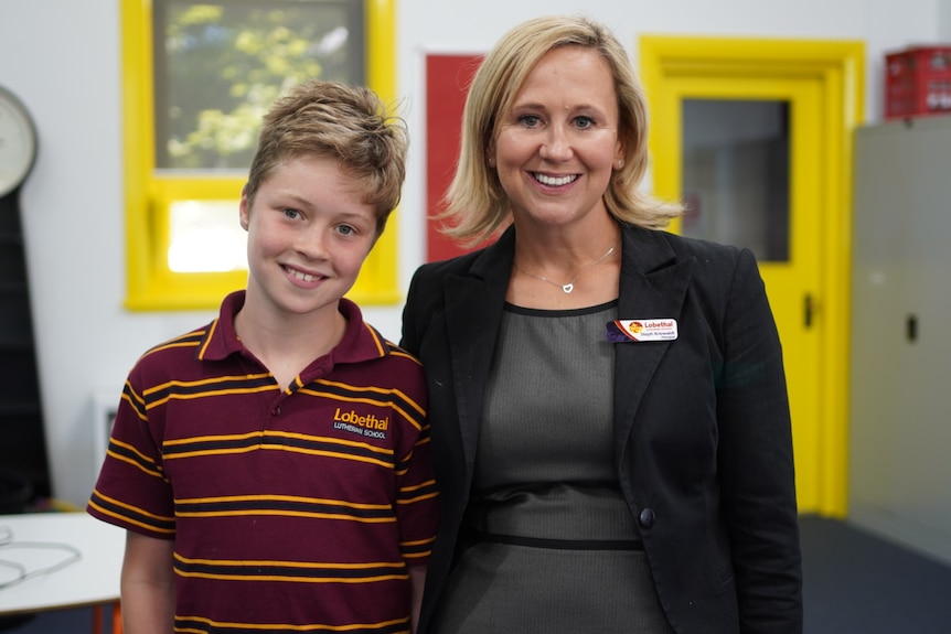 A young boy in a striped school polo shirt smiles as he stands next to his principal, a woman in a black jacket and gray dress