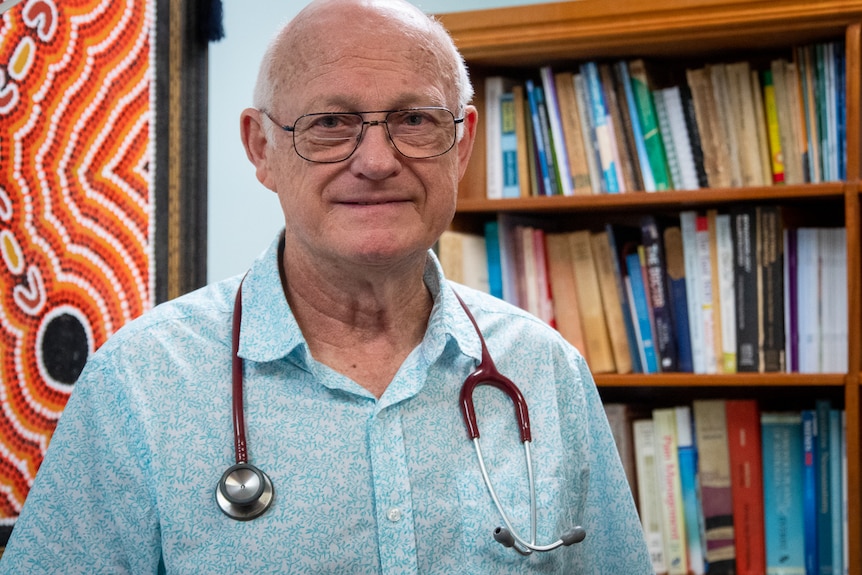 Doctor wearing a blue shirt and stethoscope, standing in front of a bookcase.