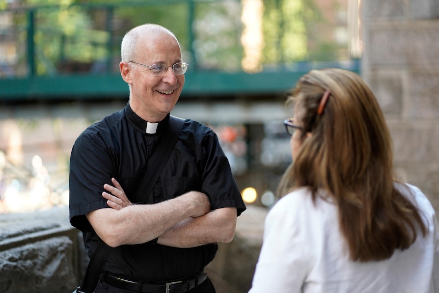 A middle-aged man in a black Catholic priest's outfit smiles with his arms folded as he speaks to a woman.