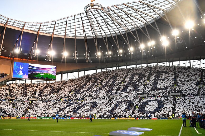 A packed crowd in a stadium hold up cards to form a black letters on a white background, but the words unclear.