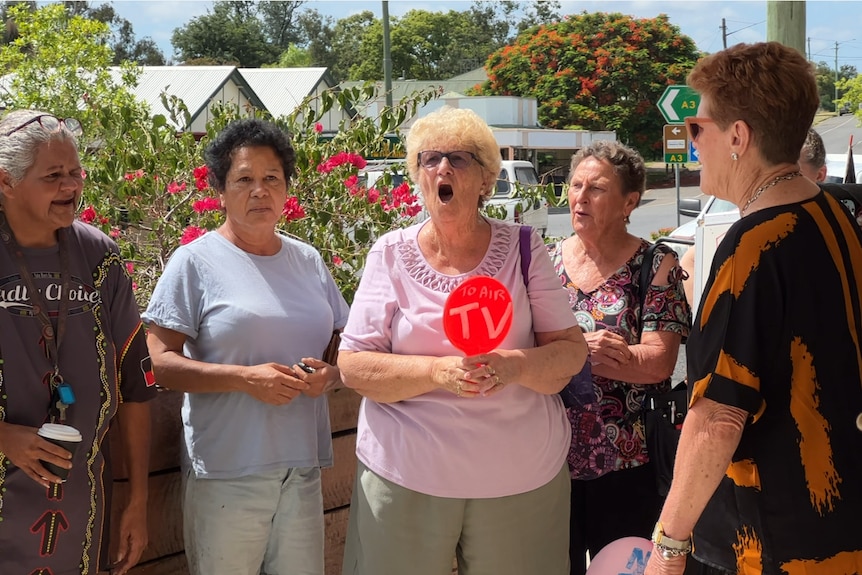 A group of women gather to protest with one shouting and holding a sign that reads, 'No TV'.