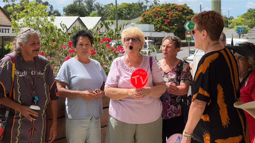 A group of women gather to protest with one shouting and holding a sign that reads, 'No TV'.