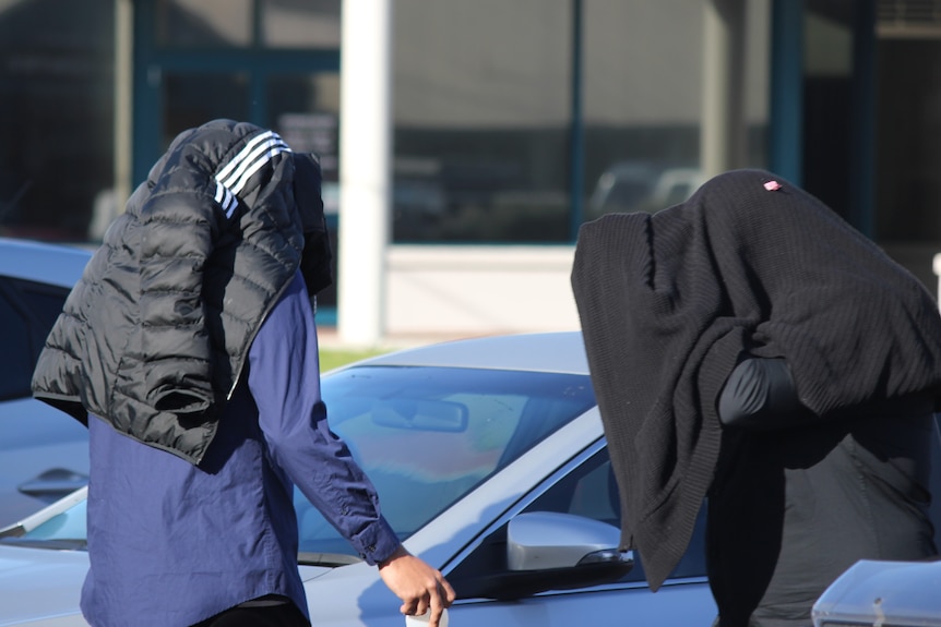 Two men with their heads covered by jackets near a car.