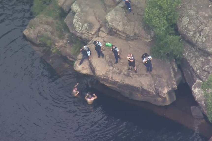 Two people can be seen in the water as police stand on a cliff looking down