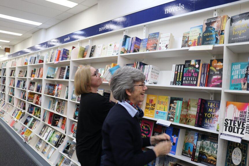 Two women with glasses and short hair are laughing and re-stacking books