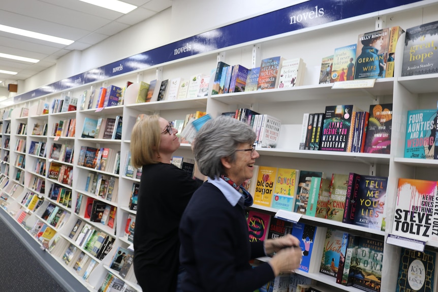 Two women wearing glasses with short hair are laughing while re-stacking books together