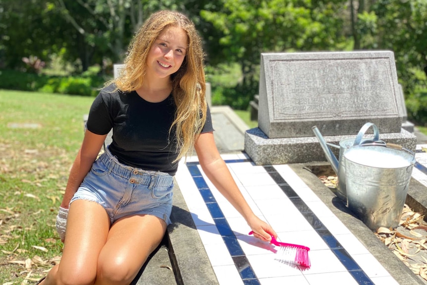 A smiling, fair-haired girl in denim shorts and a black t-shirt, sitting on a grave, holding a scrubbing brush.