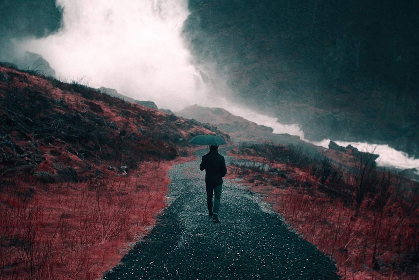 A man holds an umbrella as he walks through red and black mountains, in an apocalyptic-inspired scene.