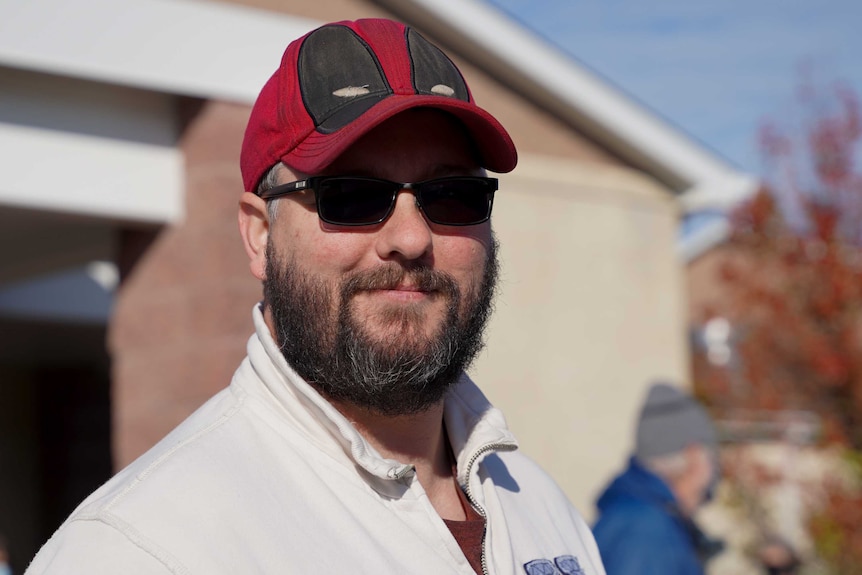 A bearded man in a cap and sunnies smiles