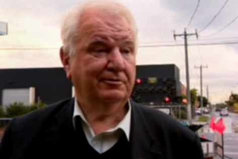 Man with white hair, collared shirt and black coat standing in suburban street.