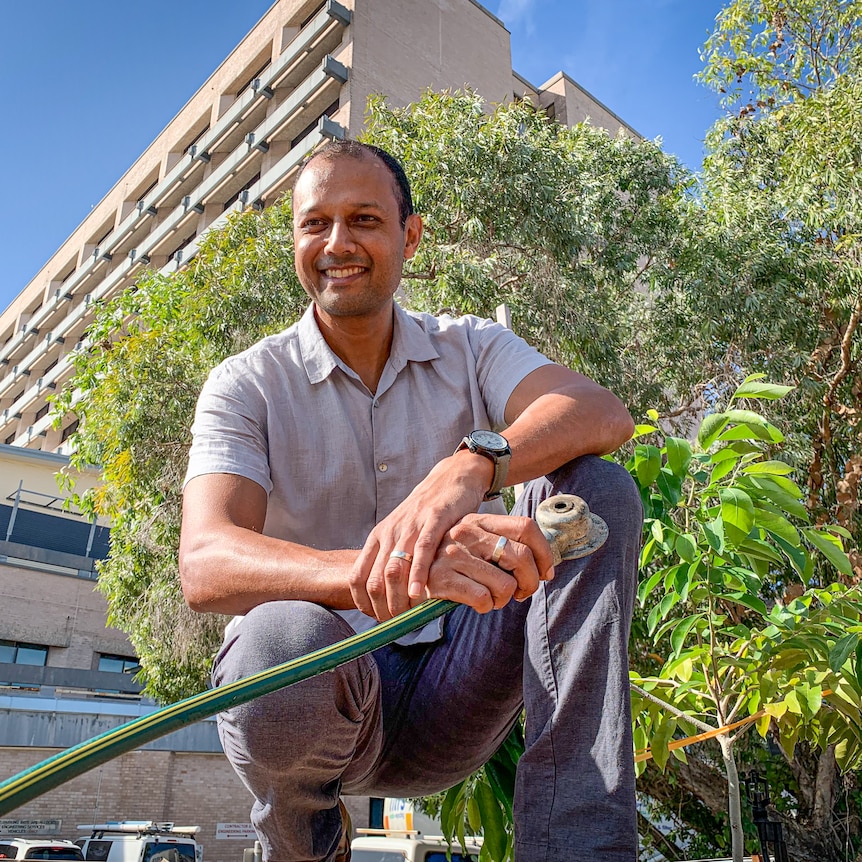 Man holding garden hose on hunkers in front of large tall building.
