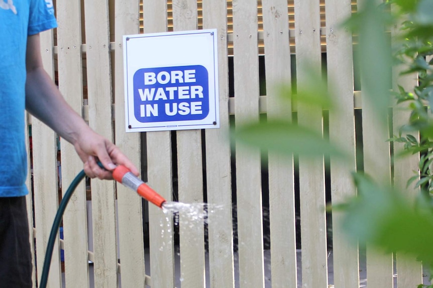 A man in a blue shirt waters a garden in front of a fence with a sign stating bore water is in use.