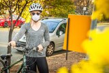 A woman is standing with her bike wearing a surgical mask near a golden wattle tree.