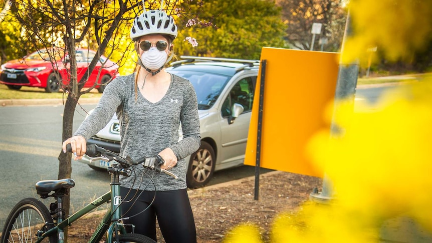 A woman is standing with her bike wearing a surgical mask near a golden wattle tree.