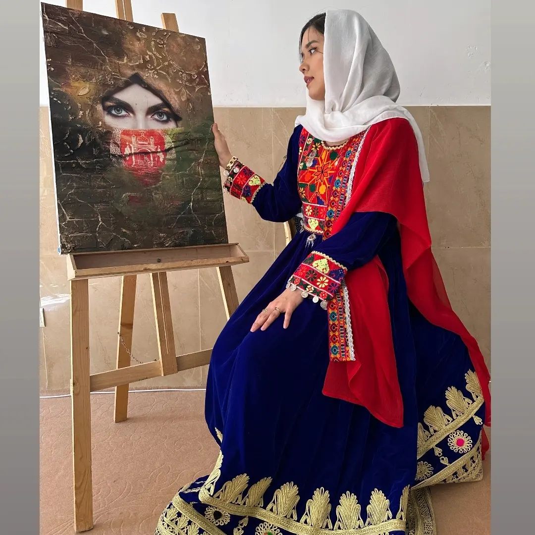 A woman wearing a white hijab and blue and red dress sitting next to an easel with a painting of a woman's face on it