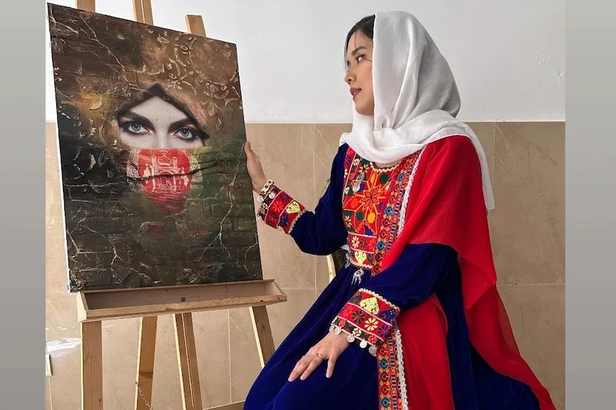 A woman wearing a white hijab and blue and red dress sitting next to an easel with a painting of a woman's face on it