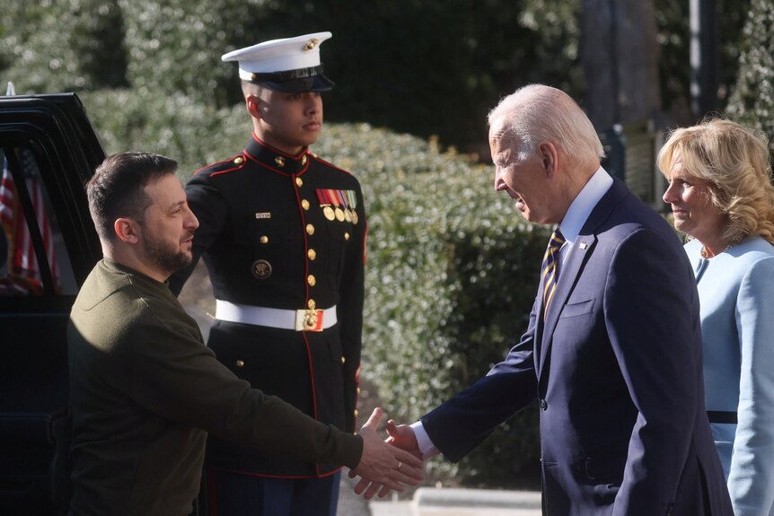 Two men shake hands in front of a guard