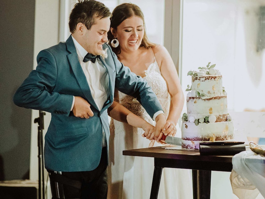 A young man and woman together cut a three-tiered wedding cake. The man has a brace on his right leg.