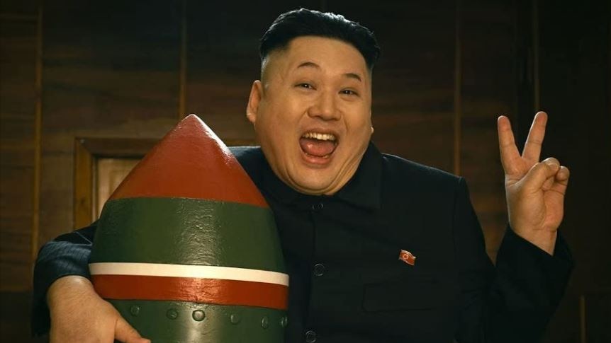 Man with close-cut black hair resembling Kim Jong Un smiles widely as he hugs a model bomb and gives peace sign with fingers.
