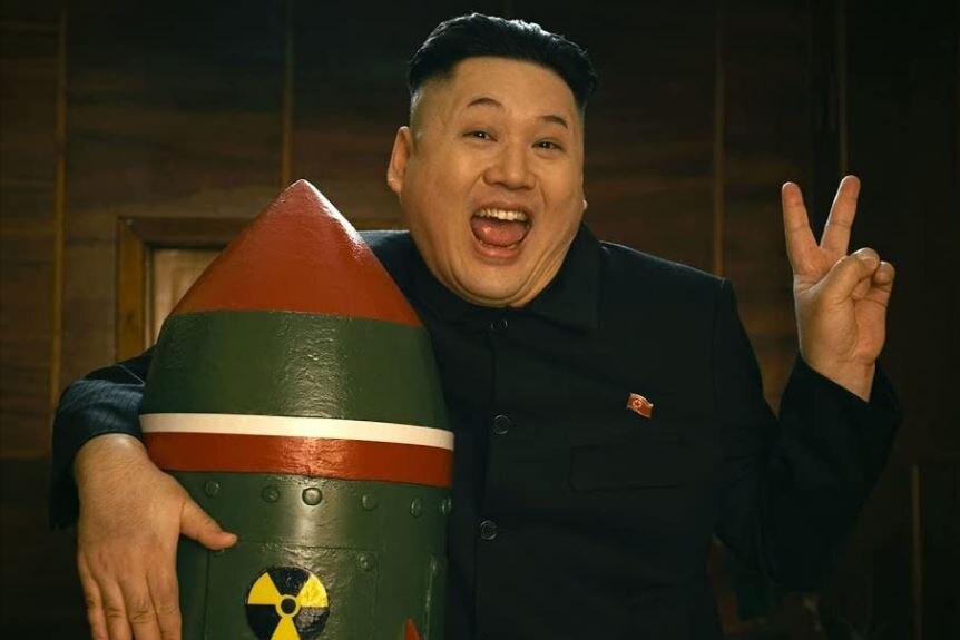 Man with close-cut black hair resembling Kim Jong Un smiles widely as he hugs a model bomb and gives peace sign with fingers.