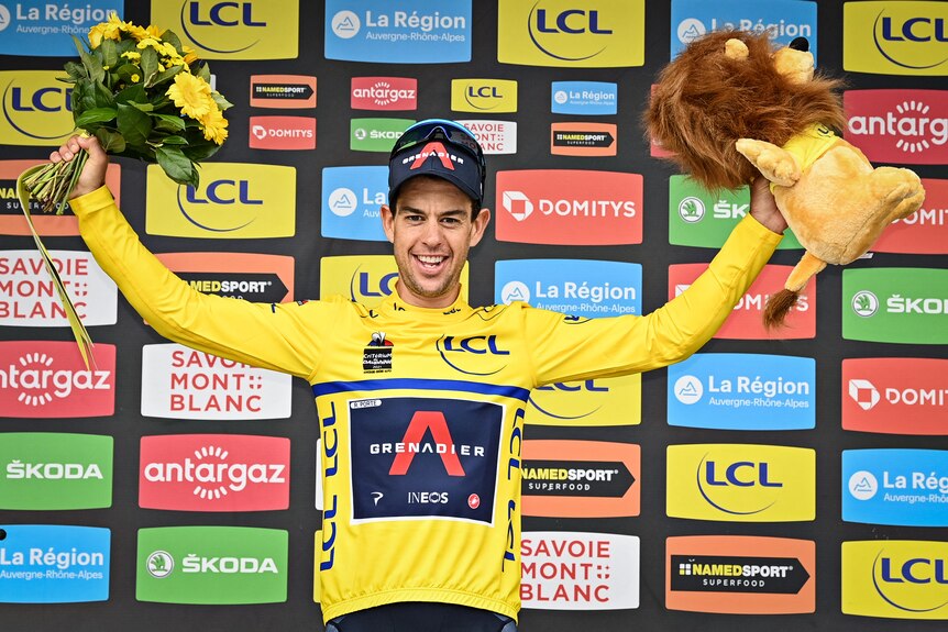 Richie Porte wears a yellow jersey and holds a bunch of flowers and a toy lion in each hand 