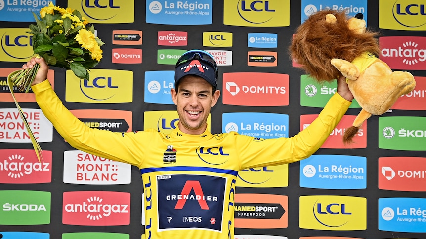 Richie Porte wears a yellow jersey and holds a bunch of flowers and a toy lion in each hand 