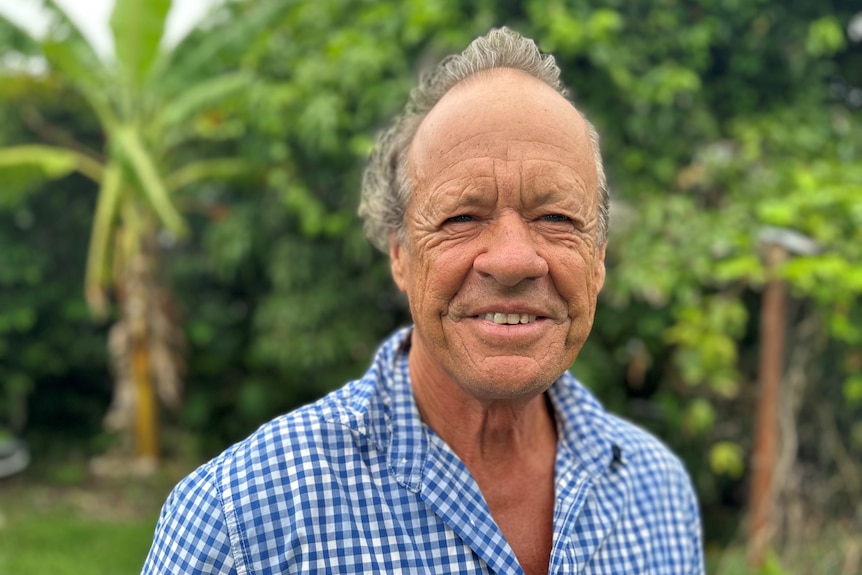 a man looks directly at a camera and smiles, he is wearing a checkered blue shirt and is outside