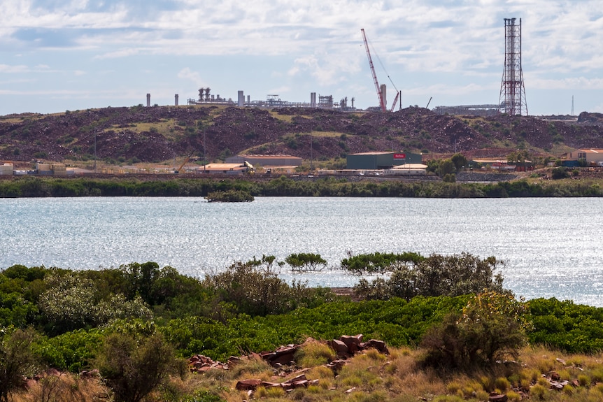 Large scale gas pumping infrastructure including towers and silos sits on a peninsula with red rock and blue ocean close by