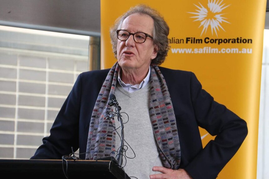 Actor Geoffrey Rush at a podium with his left hand on his hip as he speaks.