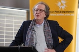 Actor Geoffrey Rush at a podium with his left hand on his hip as he speaks.