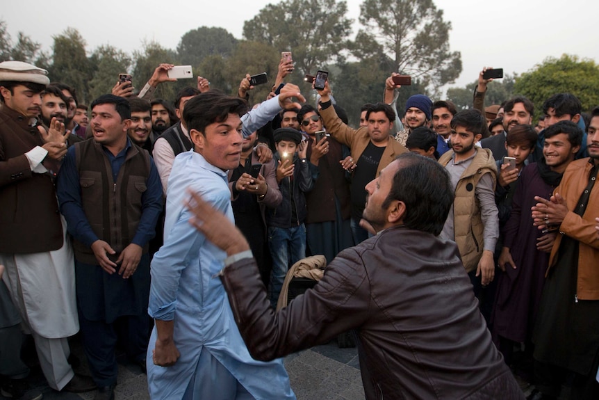 A crowd of men with smartphones watch on while contemporary and traditional clothes as two men dance.