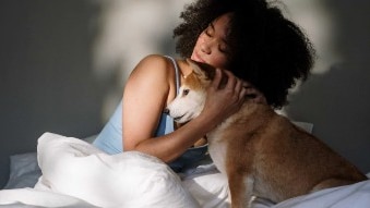 Photo of a woman with curly hair and a light blue singlet hugging a small orange and white dog