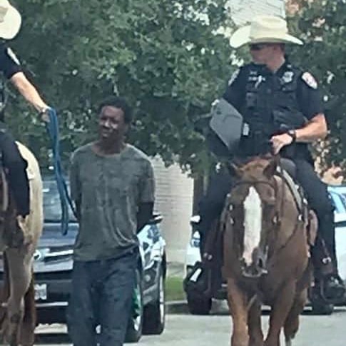 A slightly pixelated image shows two white police officers on horses leading a black man in custody on a leash.