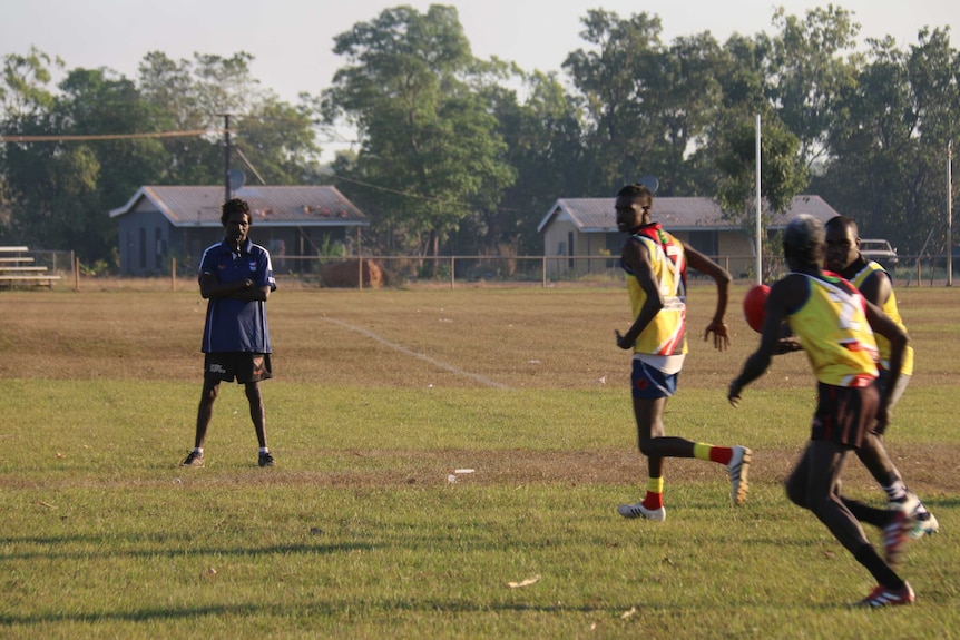 Aboriginal man in a blue shirt with a whistle in his mouth to the left and players passing a ball in a drill to the right.