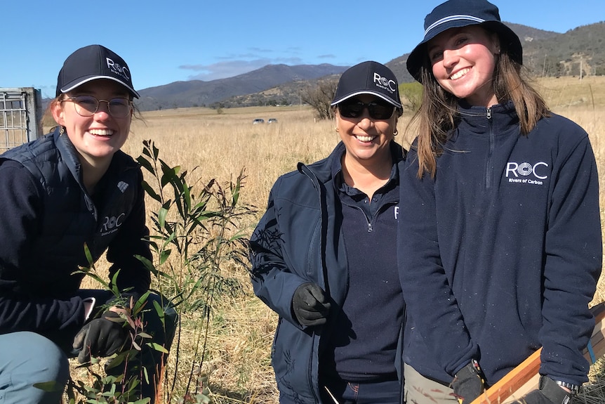 Three women smile in front of a shrub they have planted.
