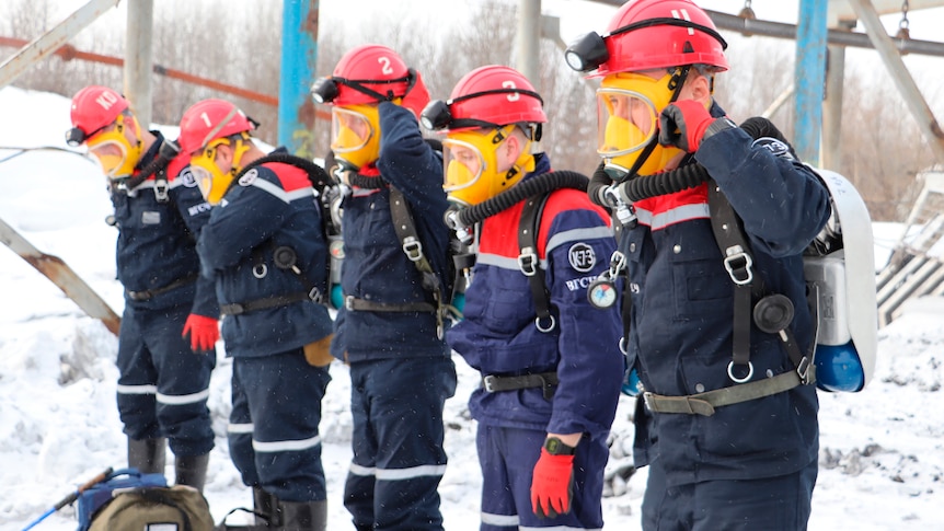 Six rescue officers wearing hard hats and breathing apparatus stand in a line in the snow. 