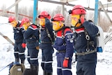 Six rescue officers wearing hard hats and breathing apparatus stand in a line in the snow. 