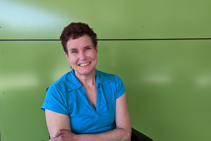 A white, middle-aged woman with short brown hair and a turquoise top smiling, and sitting in a wheelchair. 