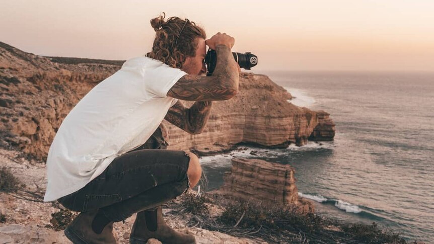 A photographer crouches on a cliff top to take a photograph.