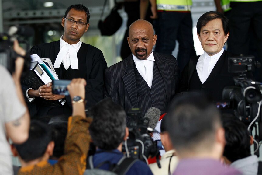 Three lawyers in robes stand on a courthouse steps talking to media.