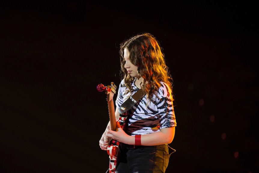 A girl with hair past her shoulders strums a dynamic electric guitar under the glow of yellow stage lights.
