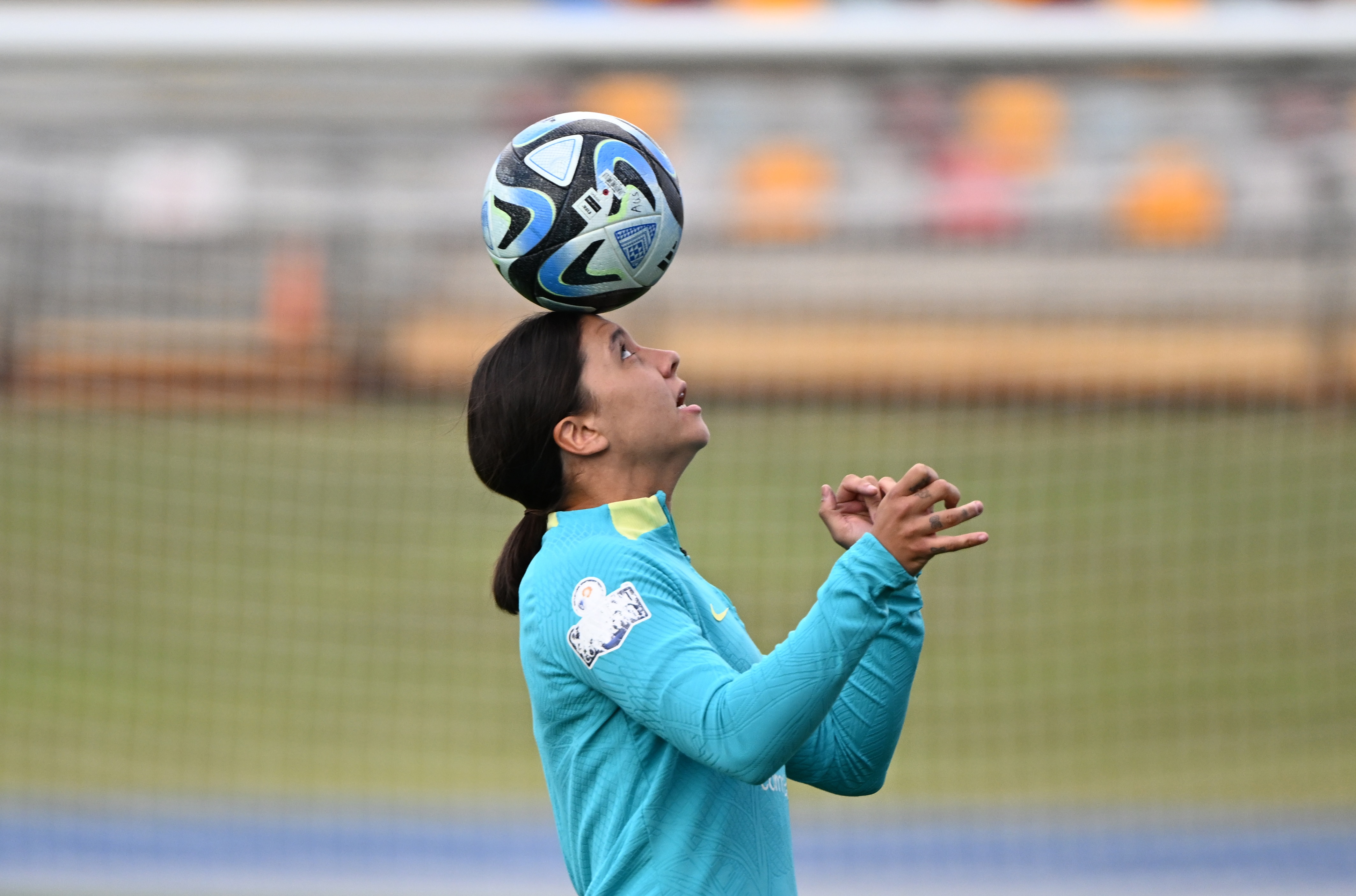 A woman in a teal jersey carefully balances a soccer ball on her head on a soccer pitch.