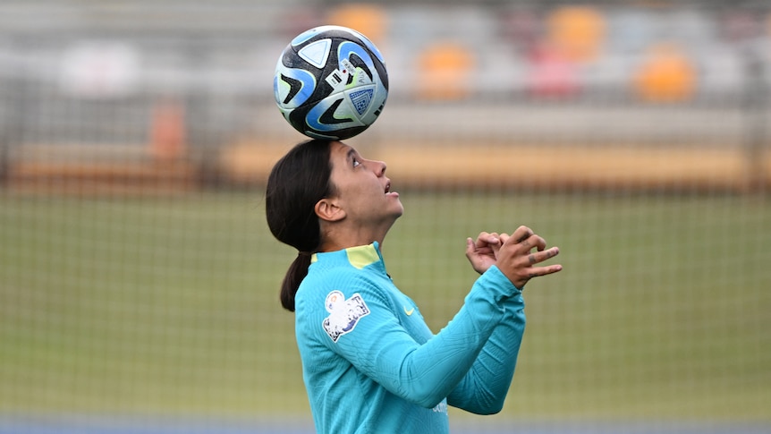 A woman in a teal jersey carefully balances a soccer ball on her head on a soccer pitch.