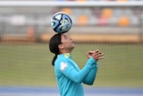 A woman in a teal jersey carefully balances a soccer ball on her head on a soccer pitch.